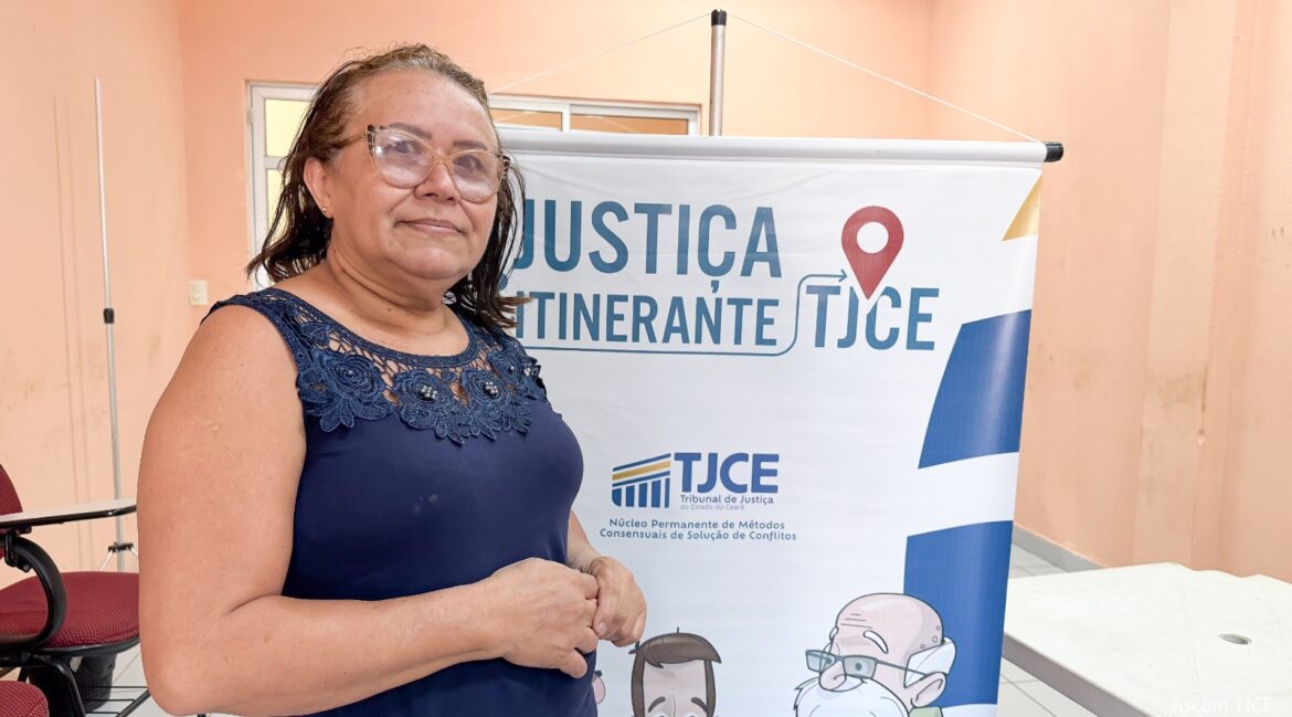 Foto de uma mulher de óculos, vestida com blusa azul, em pé, e atrás dela, um banner do Justiça Itinerante