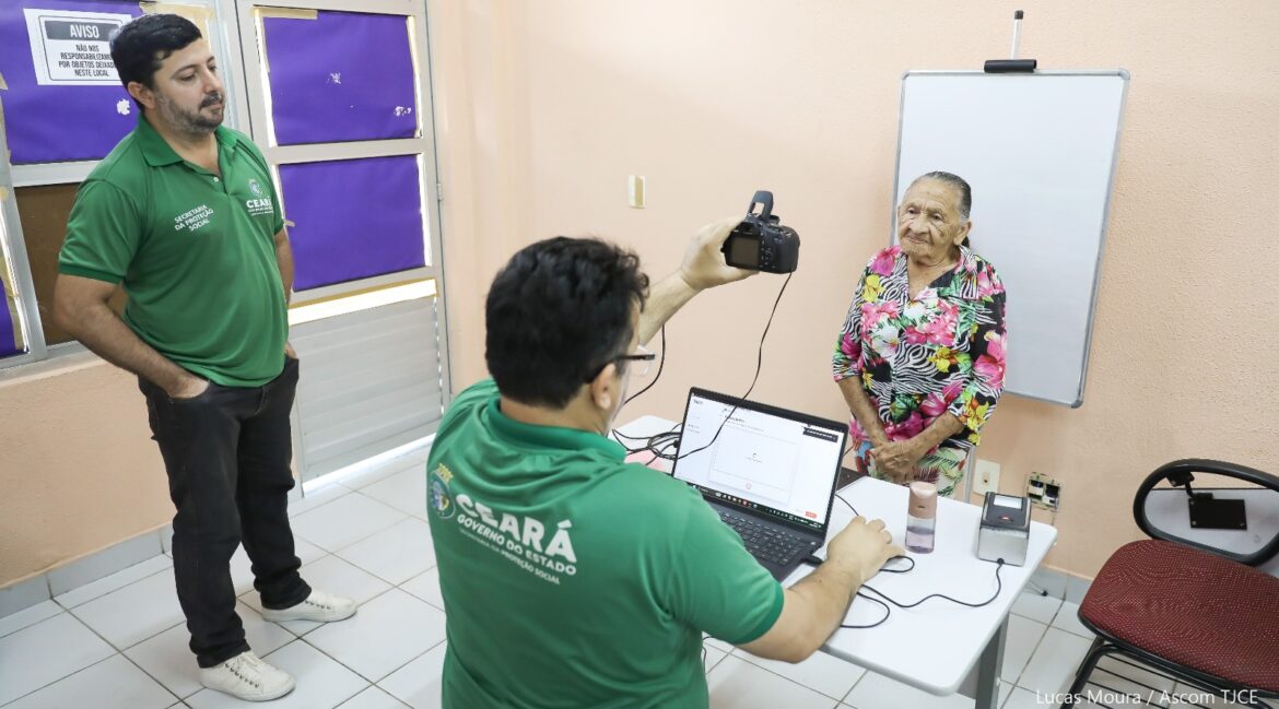 Foto que mostra dois homens e uma senhora, em uma sala. Um deles tira a foto dela, enquanto outro observa, todos estão em pé
