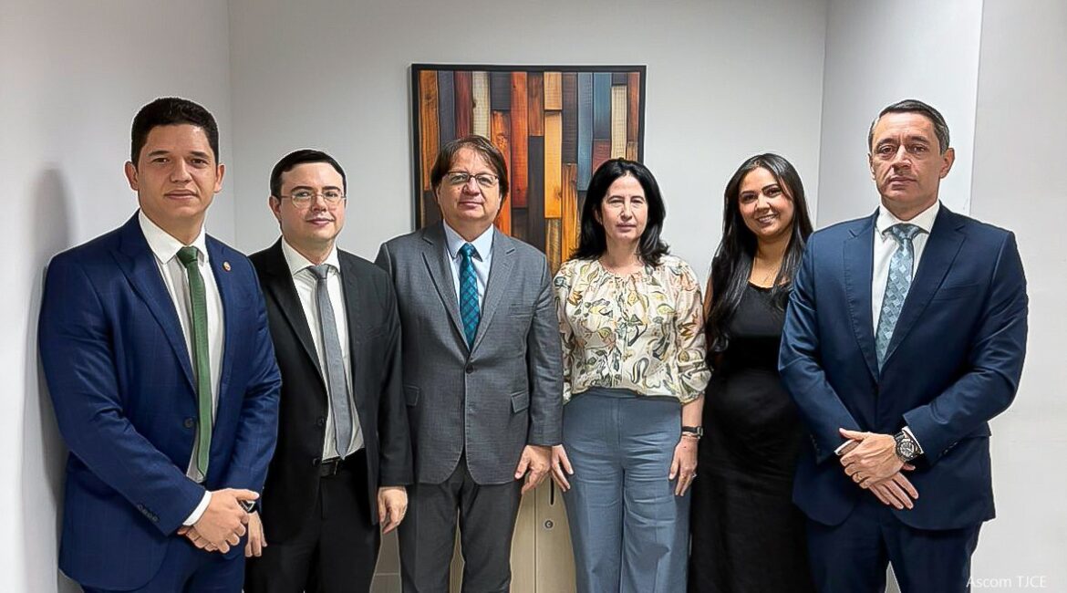 Imagem de quatro homens e duas mulheres em pé, sorrindo para foto, em uma sala