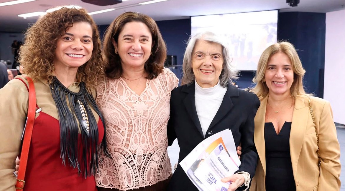 Imagem de quatro mulheres perfilhadas, em pé num auditório, sorrindo para foto