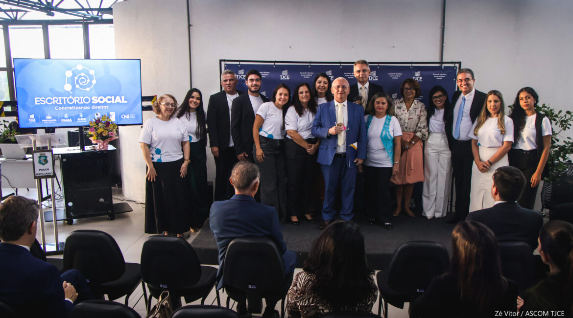 Foto em que se vê várias mulheres e homens no palco, em pé, e parte da plateia, sentada, em um grande espaço. Ao deles, um telão com o nome Escritório Social
