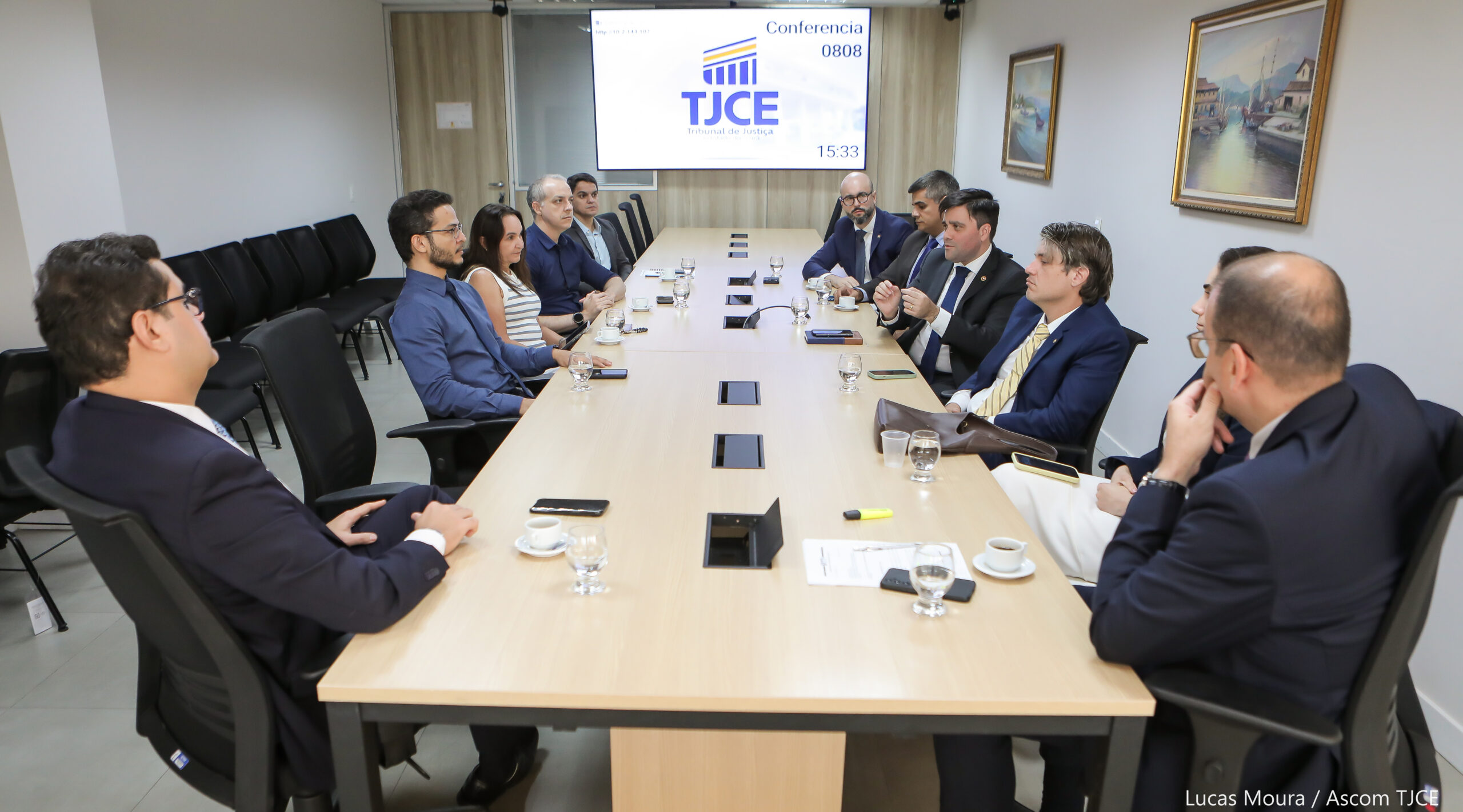 Mesa de trabalho, em que estão sentados vários homens e mulheres dialogando. À frente deles, na parede, há um grande telão com informações sobre o assunto tratado