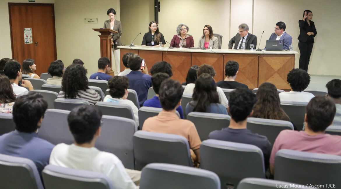 Foto geral em que se vê vários alunos e alunas de costas, na plateia, e à frente deles, uma mesa composta por dois homens e três mulheres sentadas