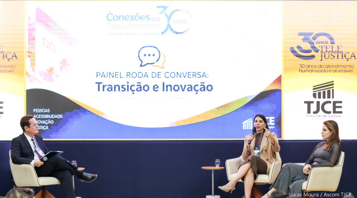 Imagem mostra duas mulheres e um homem sentados no palco, durante apresentação de uma temática