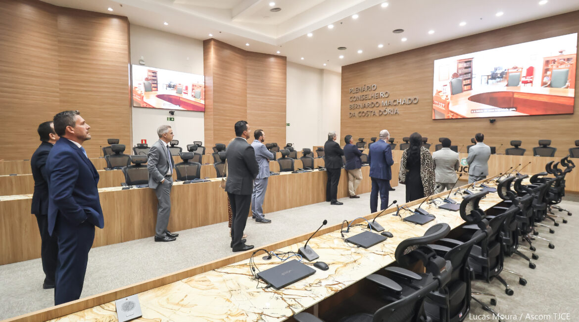 Várias pessoas de costas, observando um grande telão que está à frente deles, onde passa imagens do Memorial do Poder Judiciário