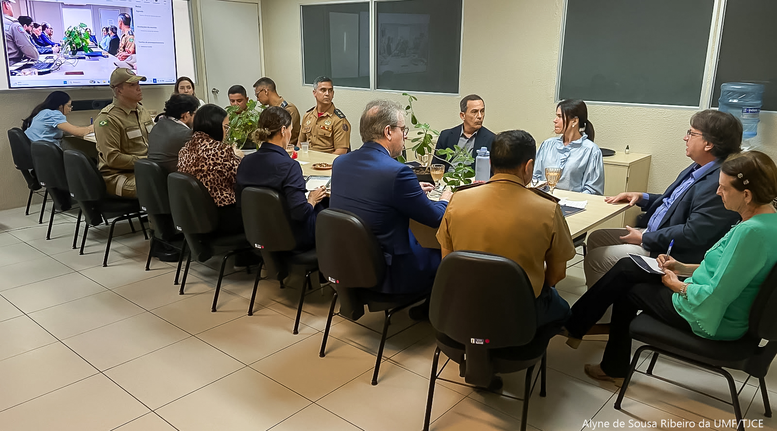 Foto de uma mesa com várias pessoas sentadas, dialogando em uma grande sala. Afixado na parede, um grande telão com imagem de outras pessoas também