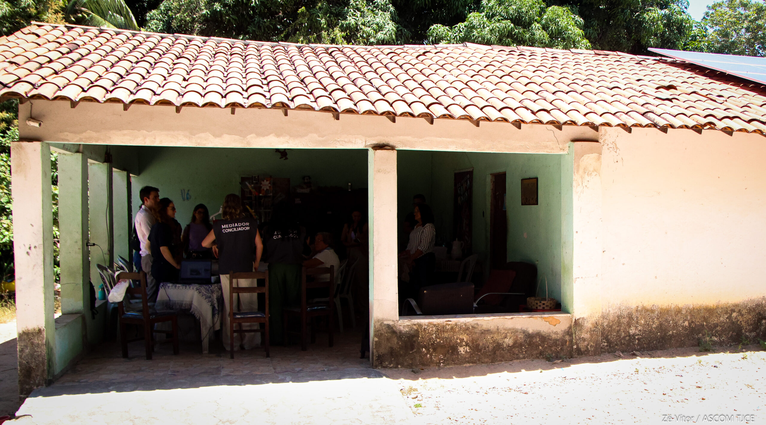 Imagem de uma casa, com varanda, em que se vê de longe, a presença de pessoas dialogando, mas que não dá pra identificá-las claramente