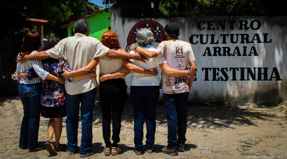 Imagem que mostra quatro pessoas abraçadas, de costas para não serem identificadas. À frente delas, uma frase grande: Centro Cultural Arraiá Testinha, escrita com tinta preta na parede branca, ao ar livre