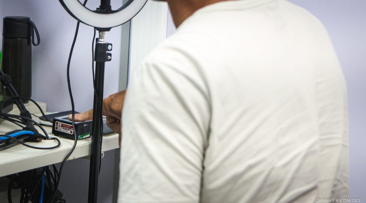 A imgam mostra um equipamento de luz de anel com suporte de tripé e um leitor biométrico de impressões digitais sobre a bancada. À frente do equipamento, se vê as costas de um homem, que tem o dedo em um aparelho para coleta de impressão