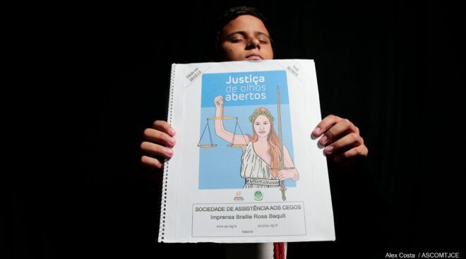 Foto que mostra um garoto cego, exibindo uma cartilha em Braille, onde está escrito: Justiça de Olhos Abertos com a deusa themis sem a venda dos olhos