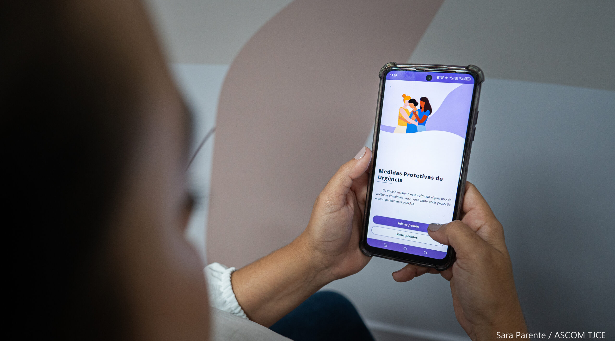 A imagem mostra uma mulher de costas, com destaque para um celular na mão, que mostra a tela do aplicativo TJCE Mobile, para a solicitação de Medidas Protetivas de Urgência.