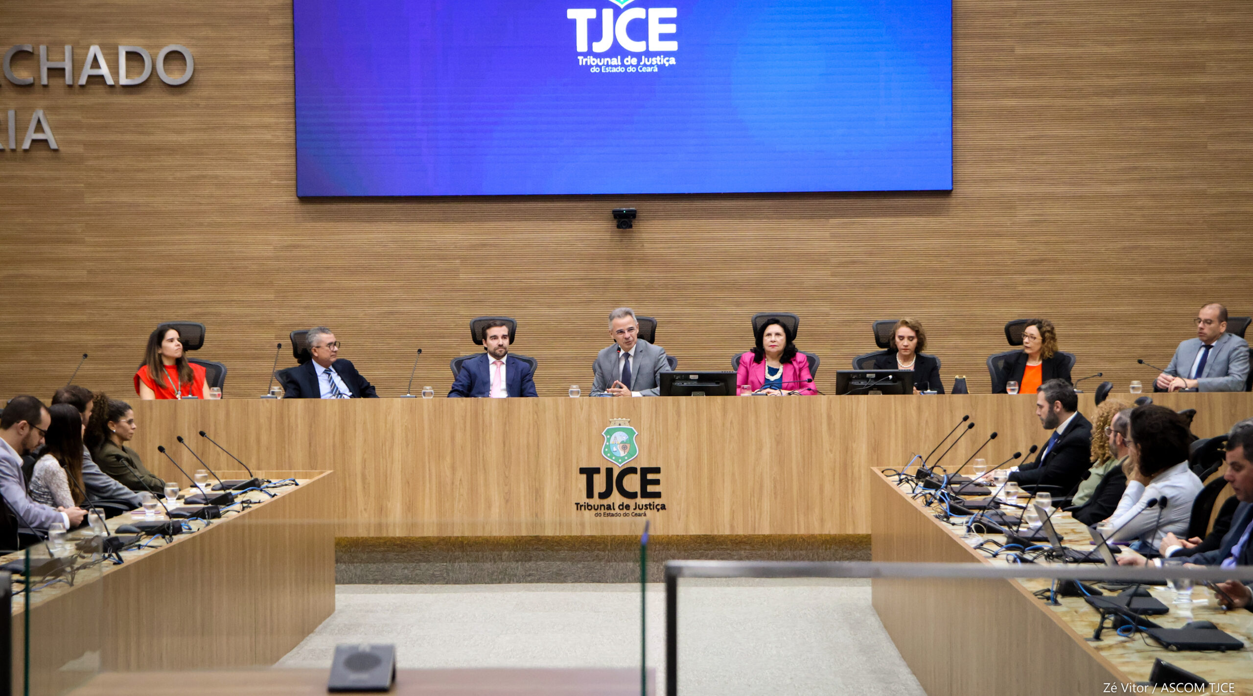 Pessoas sentadas por trás de bancadas na cor madeira. Ao fundo, telão há um telão com imagem na cor azul e as palavra: TJCE - Tribunal de Justiça do Ceará e o brasão do Estado do Ceará.