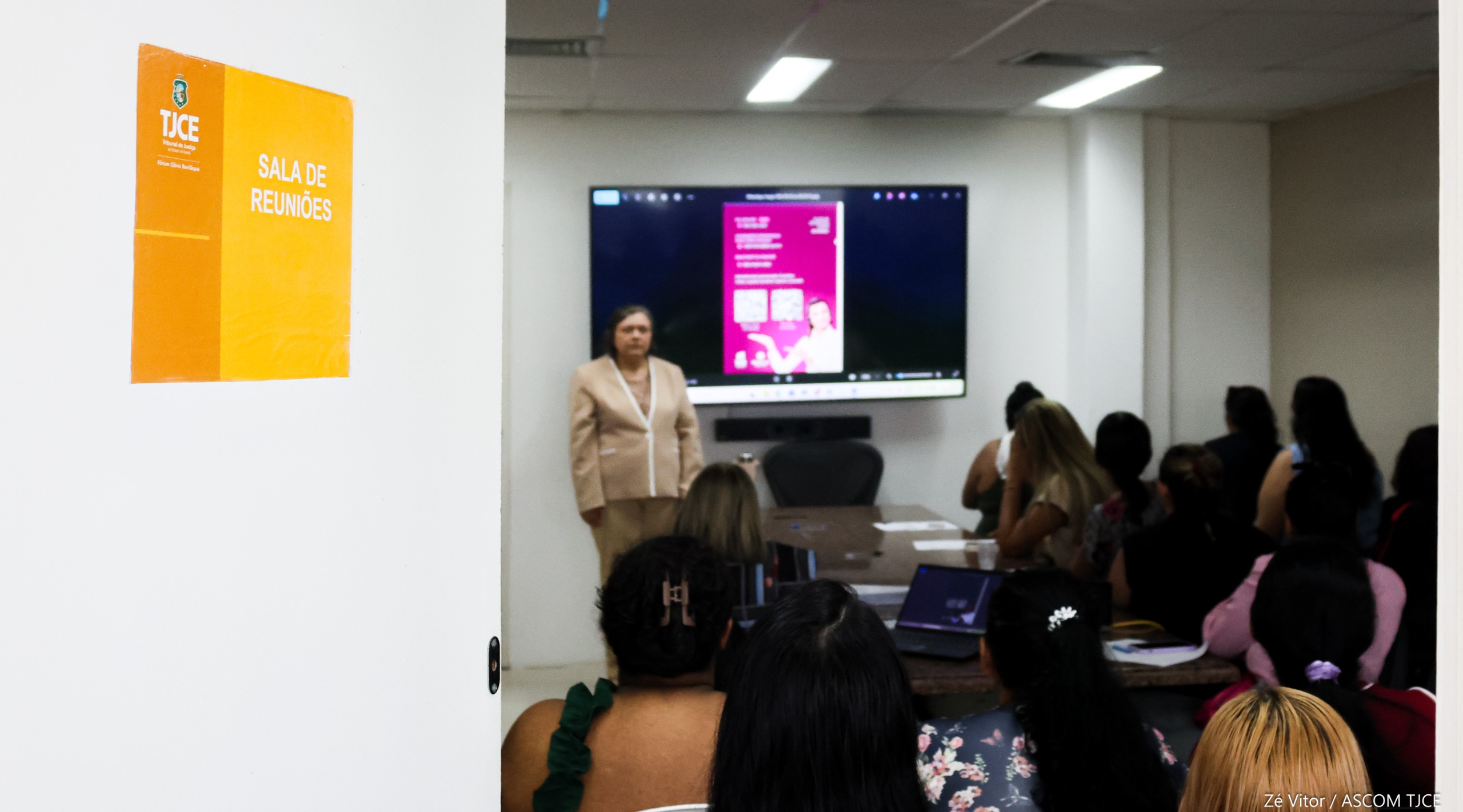 Foto geral m que se vê ao fundo, uma mulher falando, para 29 outras mulheres sentadas, em uma sala