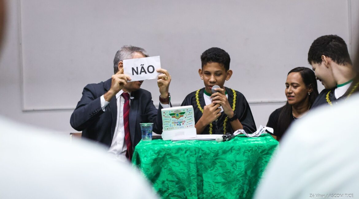 A foto mostra um juiz de terno e gravata vermelha, segurando um papel com a palavra "NÃO" escrita em letras maiúsculas. Ao dele, o jovem arthur está com um microfone na mão, dando o resultado do júri. Ao lado dele, uma professora está sentada auxiliando ele.