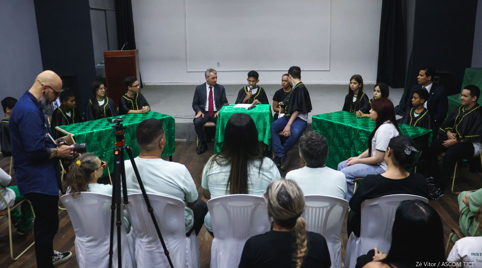 Foto geral do espaço onde foi realizado o júri simulado. Nele, se vê várias pessoas na plateia e a composição dos jovens simulando o júri, sentados à direita, à esquerda e ao centro do auditório