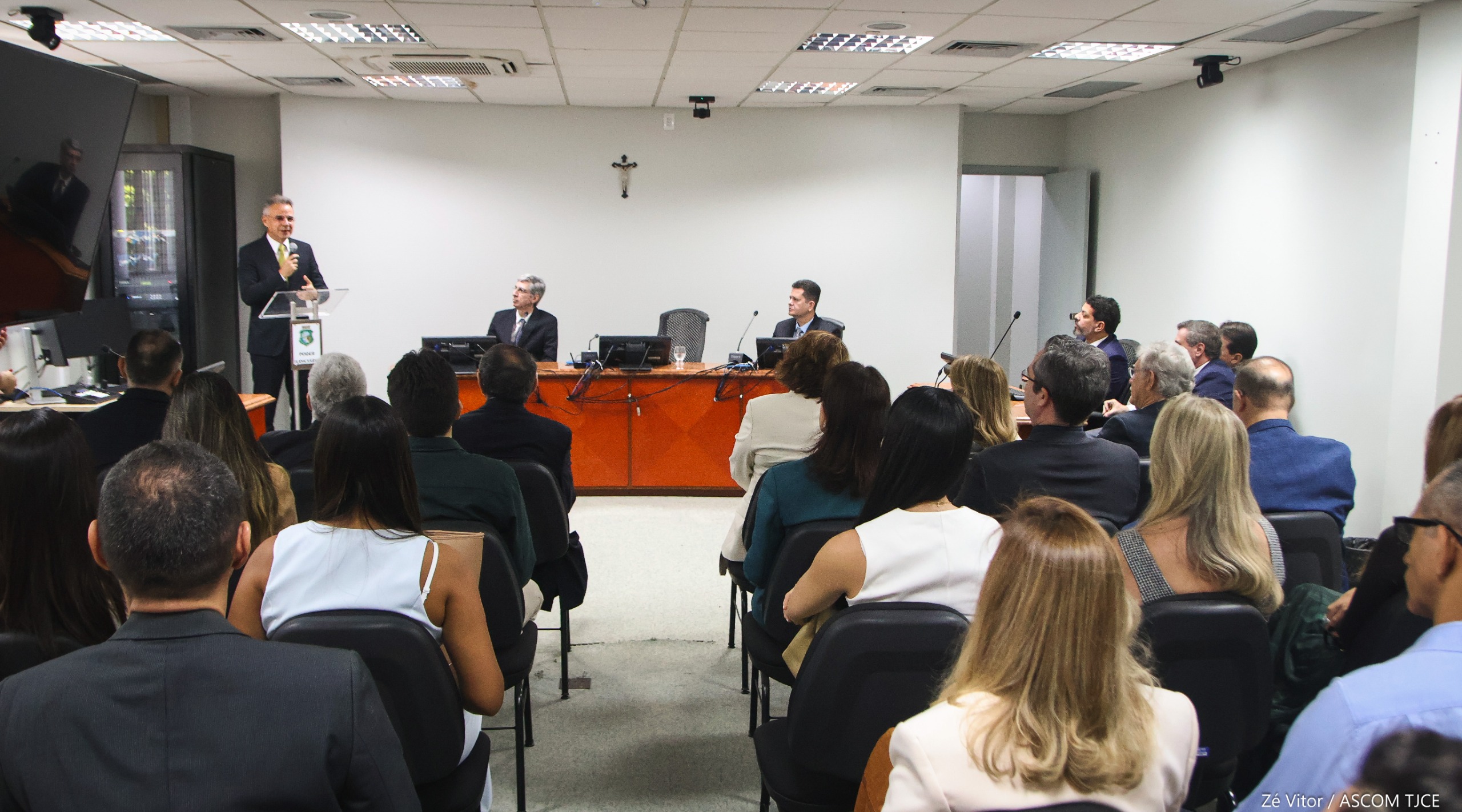 Foto de um homem falando ao microfone no palco e, à frente dele, várias pessoas sentadas no auditório. Ao dele, dois homens sentados observando-o falar.