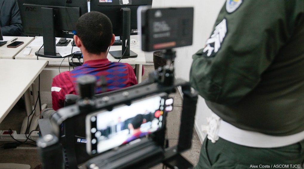 Imagem de um homem de costas sentado. À frente dele, monitores de computador. Atrás, uma tela de celular em um suporte, com a mesma imagem dele sendo filmada, e um policial com fardamento verde da Polícia Militar, não identificado, enquadrado na cintura pela lateral.
