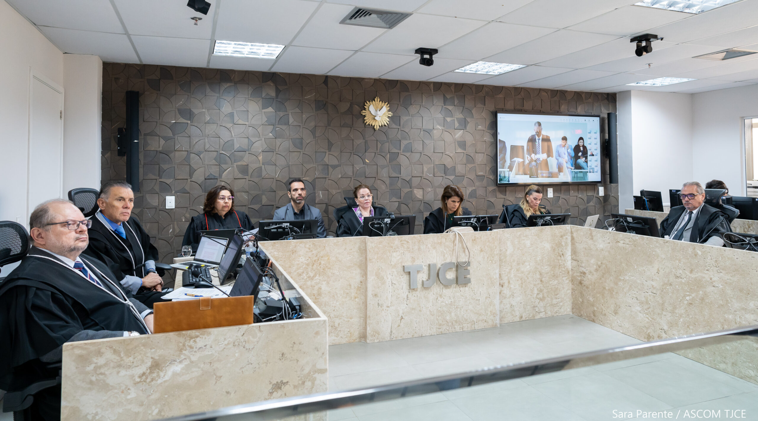 Foto de uma sessão de julgamento, onde se vê várias pessoas sentadas em uma bancada de trabalho, analisando processos