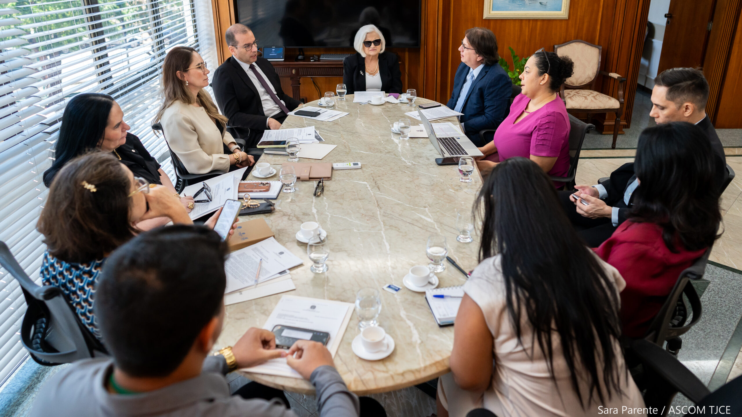 Foto com homens e mulheres sentados em volta de uma mesa de trabalho dialogando, em uma sala