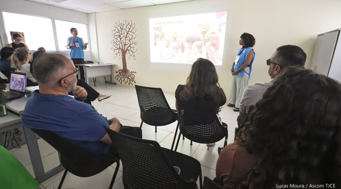 Foto de uma sala ampla, em que se vê duas pessoas à frente dando explicações sobre uma temática, e várias pessoas sentadas ouvindo, atentamente