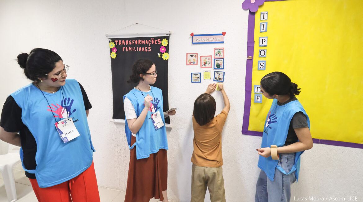 A foto mostra três profissionais que cuidam das crianças e adolescentes, observando uma criança fazer uma colagem na parede. A criança está de costas, em uma sala