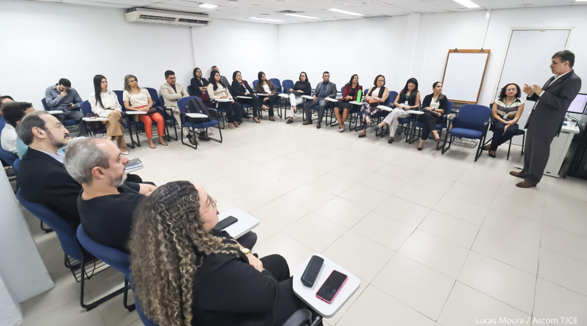 Foto de várias pessoas sentadas em cadeiras, formando um círculo e, ao centro, um homem de paletó dando explicações