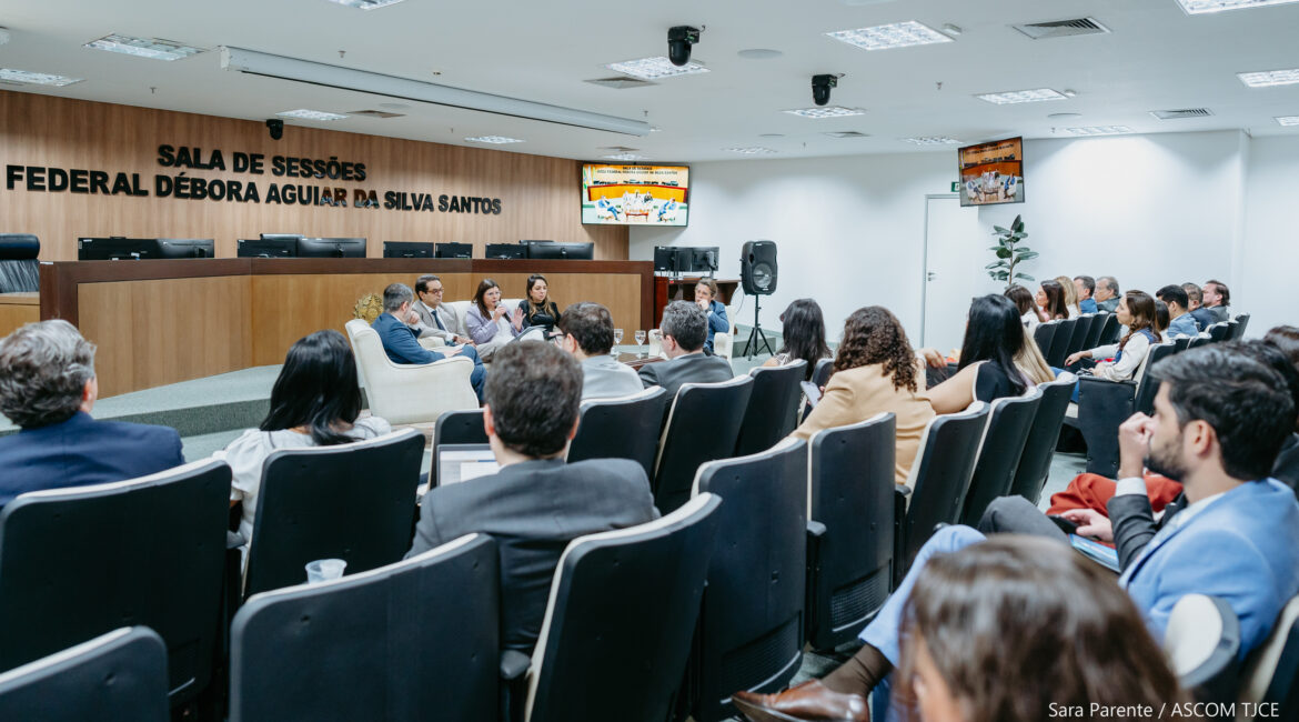 Foto de homens e mulheres à frente da plateia, sentados, onde se vê uma mulher com microfone na mão, falando ao público, que também sentado ouvindo-a