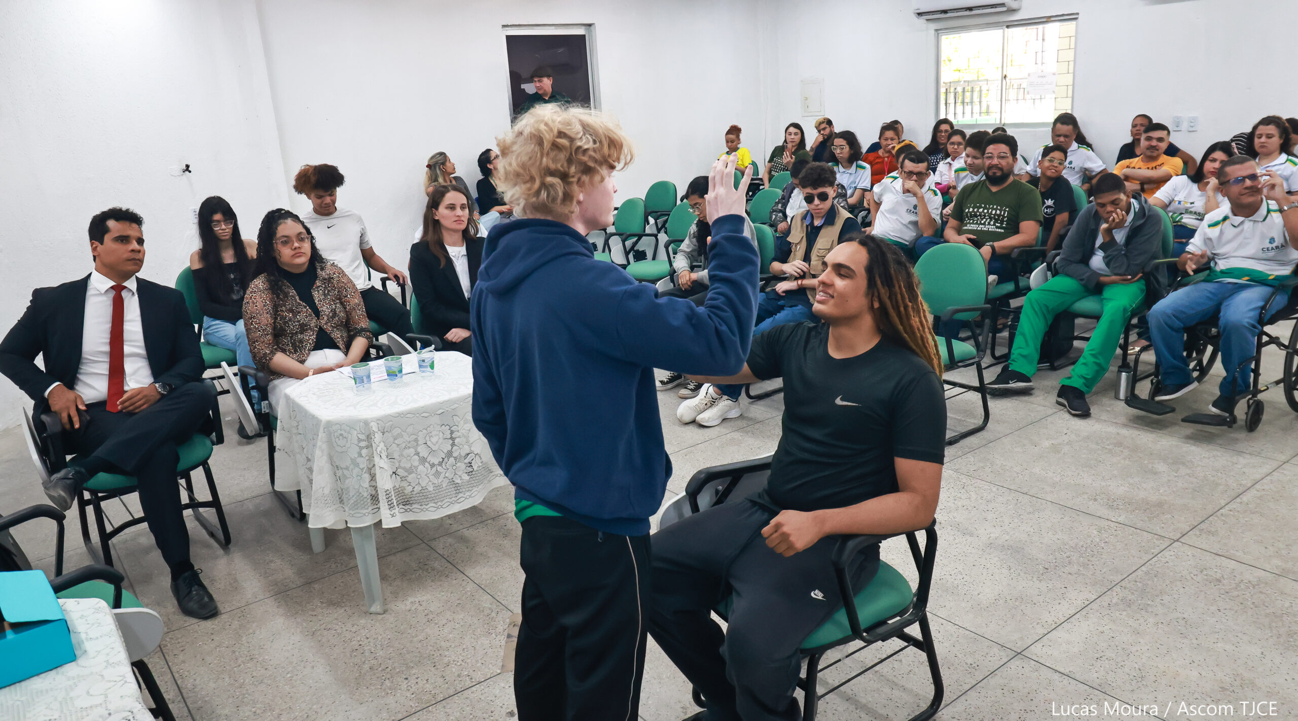 Foto geral da sessão do júri, em destaque um adolescente que está em pé com a mão erguida sobre na frente de outro jovem que está sentado, no momento do juramento da palavra de que dirá somente a verdade