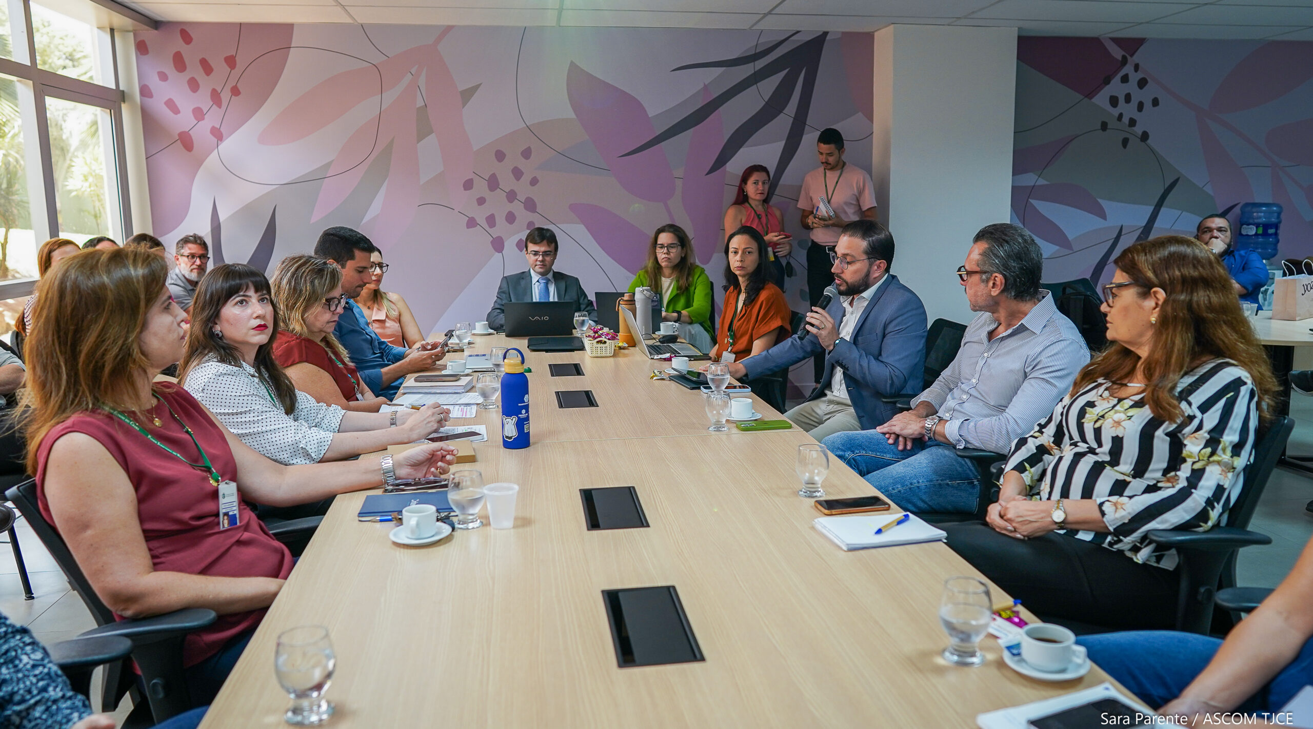Foto com vários homens e mulheres sentados, ao redor de uma grande mesa, dialogando, em uma ampla sala
