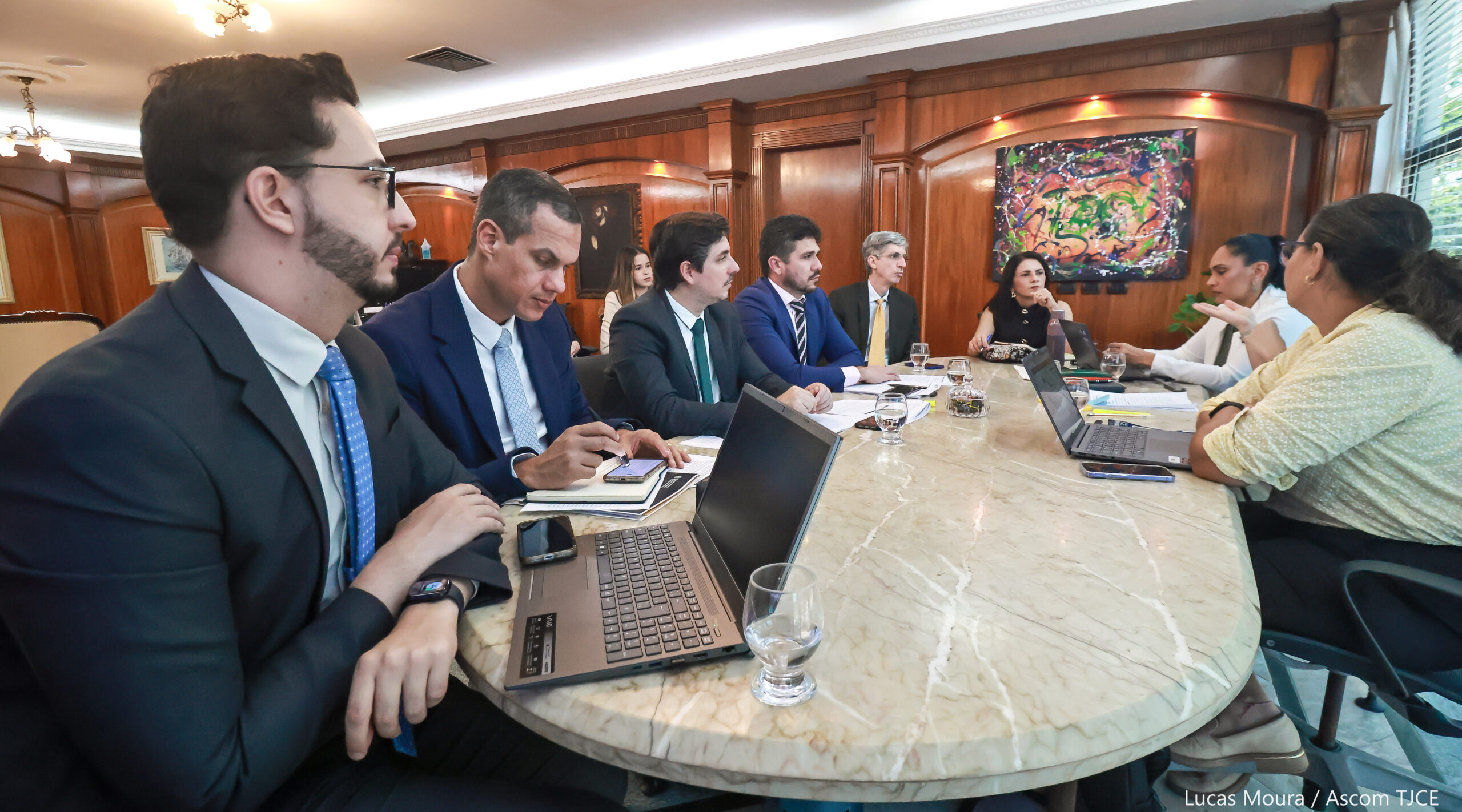 Vários homens e mulheres sentados ao redor de uma mesa, dialogando, em uma ampla sala