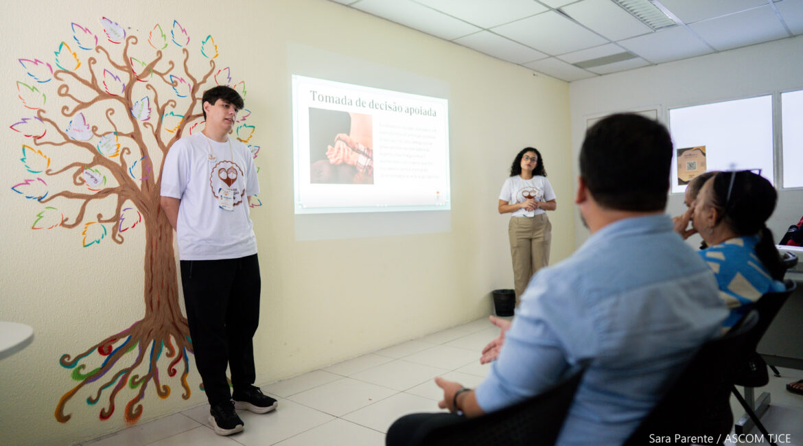 Foto de duas pessoas dando as explicações, em uma sala, em pé. Elas estão à frente de várias pessoas sentada que estão ouvindo atentamente