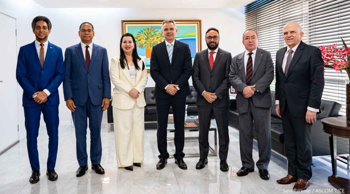 Imagem de seis homens e uma mulher, em pé, sorrindo para foto, em uma sala