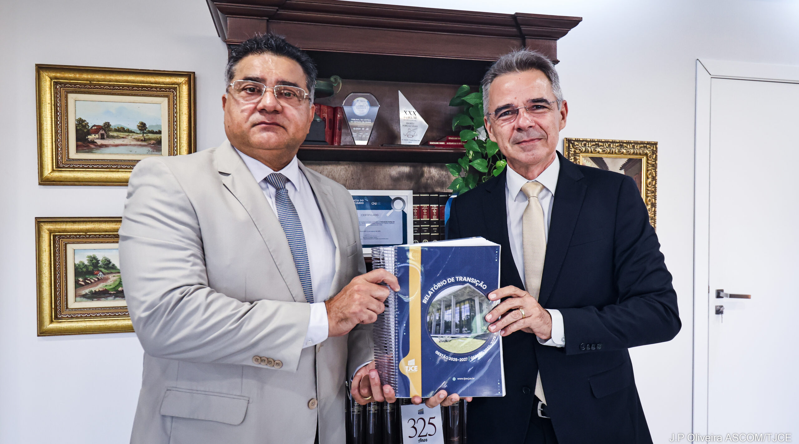 Imagem de dois homens, posando para foto, ambos segurando um documento, em uma sala