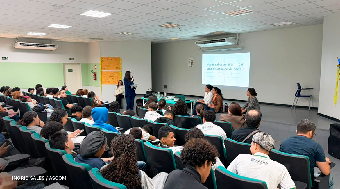 Foto de uma mulher falando ao microfone e, à frente dela, vários jovens na plateia, sentados, ouvindo atentamente. Na parede, um grande telão com a frase: Vocês saberiam identificar uma situação de violência?