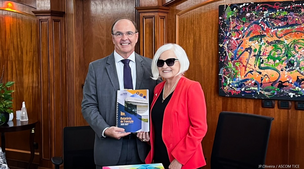 Imagem de um homem e uma mulher, juntos, sorrindo para foto, em pé, exibindo um documento, em uma sala