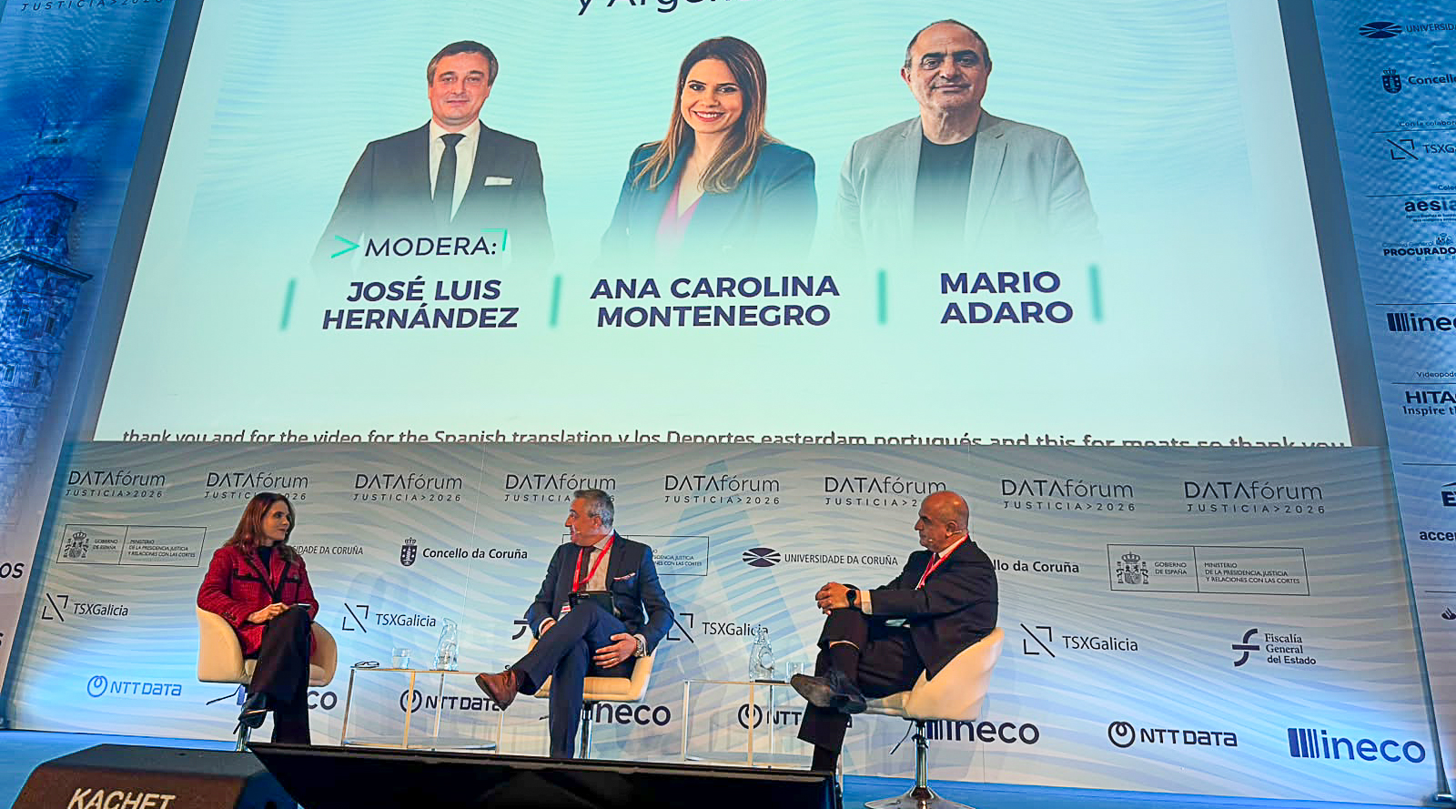 Dois homens e uma mulher, dialogando em um palco, e, ao fundo deles, um grande telão projetado na parece, com a imagem deles mesmos
