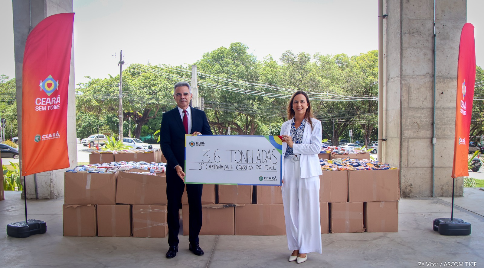 Foto de uma mulher e um homem, exibindo o cartaz em que se lê: 3,6 toneladas de alimentos, 3ª Caminhada do TJCE. Atrás deles, estão empiladas várias caixas de alimentos, em um ambiente ao ar livre.