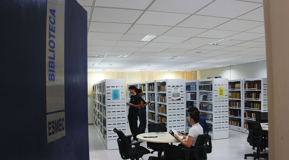Foto geral de uma biblioteca onde se vê duas pessoas folheando livros em pé, ao fundo, e uma mulher sentada numa mesa, mais em destaque